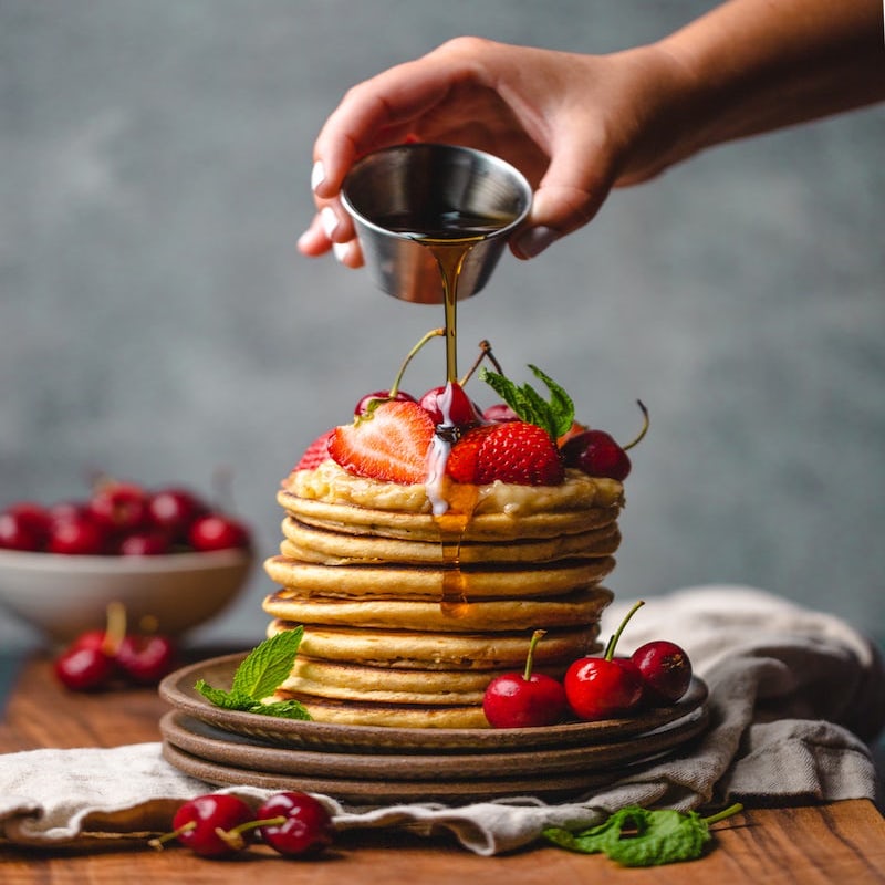 stack of pancakes with hand pouring syrup from metal pot
