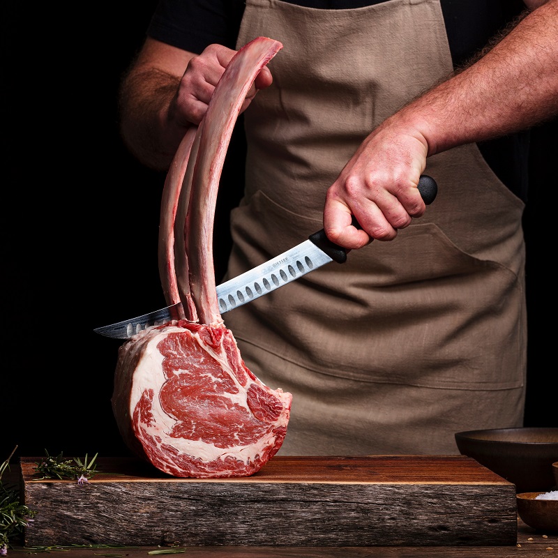 man chops a tomahawk steak in a brown apron with a large knife