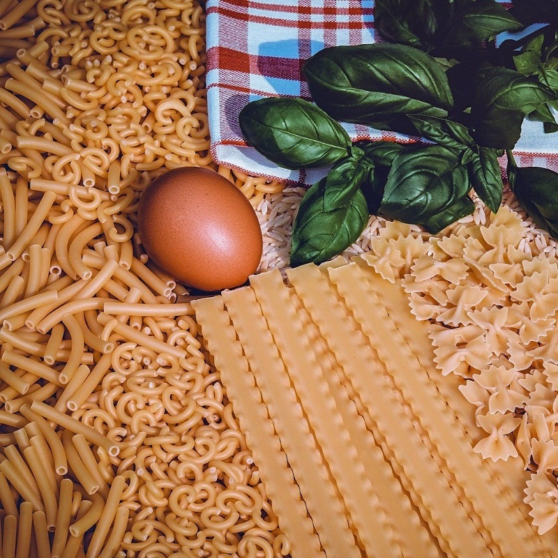 various pasta shapes laid out on a table with a checked tablecloth and basil