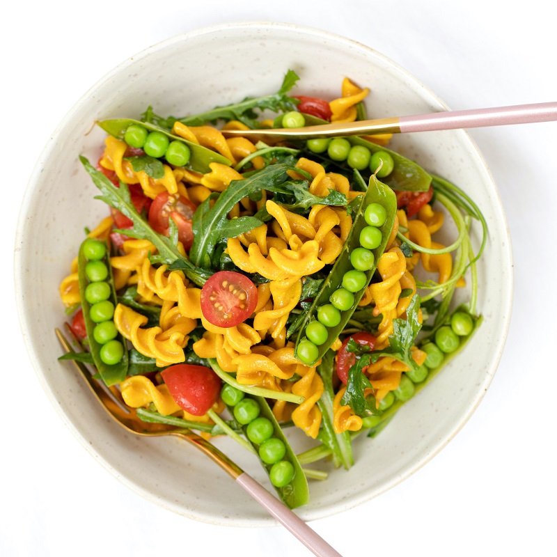 healthy pasta salad in a white bowl against a white background with tomatoes and peas
