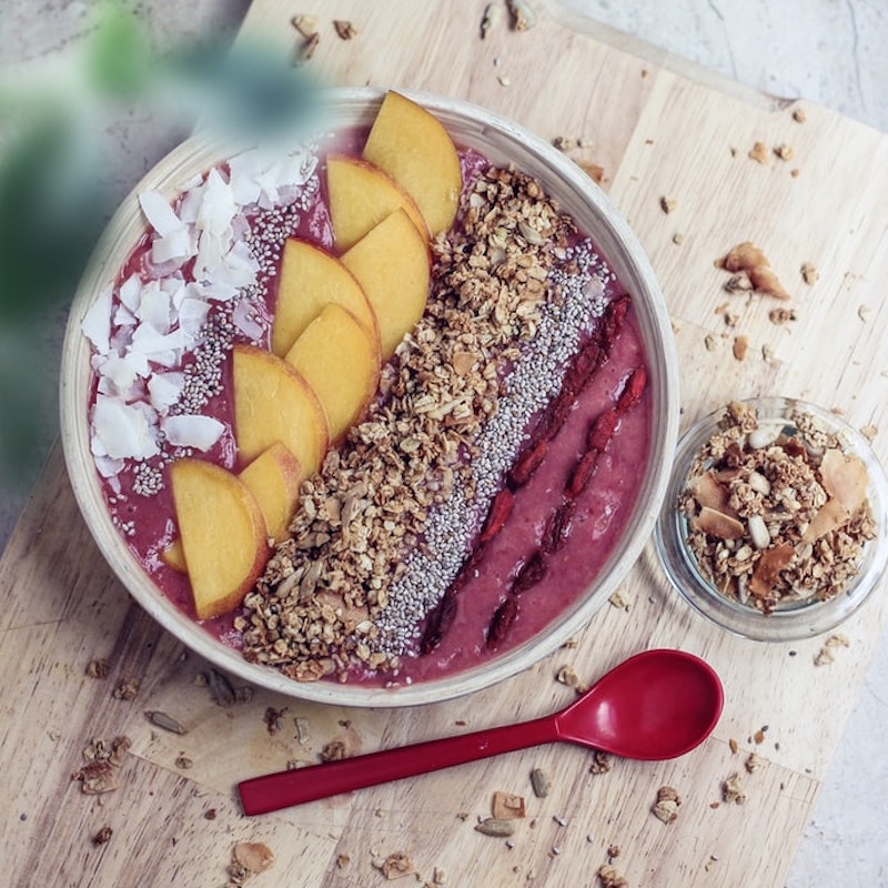 pink smoothie bowl with granola, coconut and peach arranged vertically